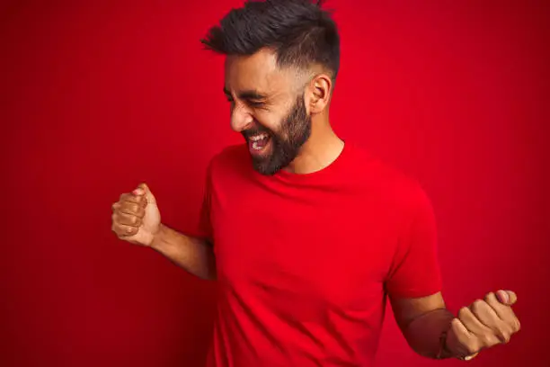 Photo of Young handsome indian man wearing t-shirt over isolated red background very happy and excited doing winner gesture with arms raised, smiling and screaming for success. Celebration concept.