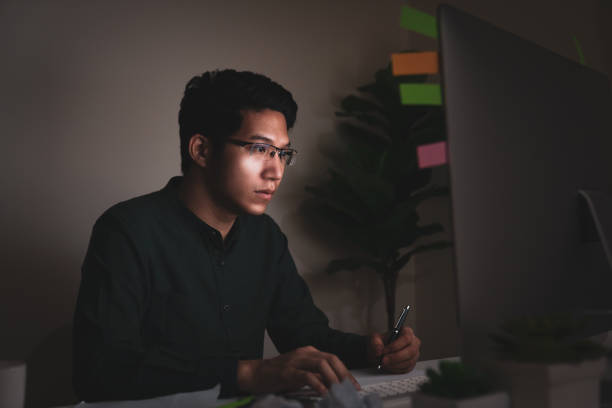 istock Attractive young asian man sitting on desk table looking at laptop computer in dark late night working feeling serious thinking and determinated at home office in work hard or work load job concept. 1167771778