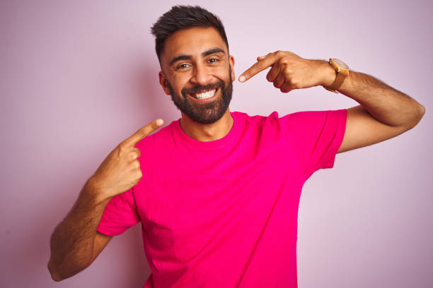 молодой индийский мужчина в футболке, стоящей над изолированным розовым фоном, улыбается веселым показом и указывает пальцами зубы и рот. к - dentist asian ethnicity portrait male стоковые фото и из�ображения