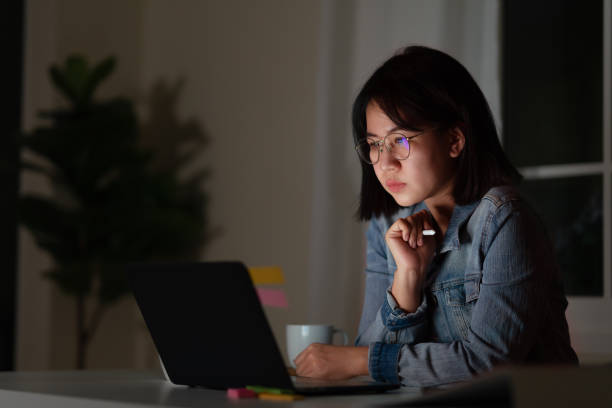 candid of young asian female student sitting on desk with smart digital gadget looking at notebook working at late night with project research, graphic designer or programmer concept. - promotor inmobiliario fotografías e imágenes de stock