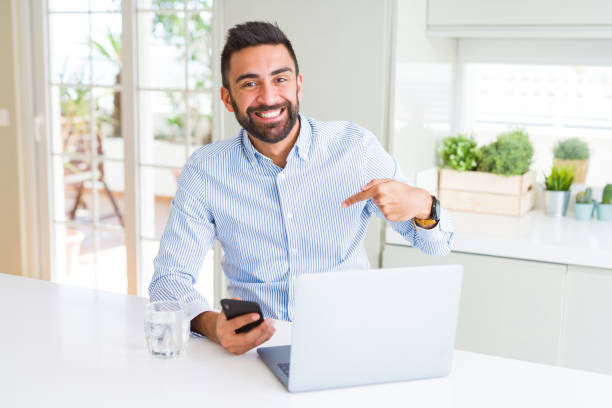 istock Handsome hispanic business man using laptop and smartphone with surprise face pointing finger to himself 1167773348