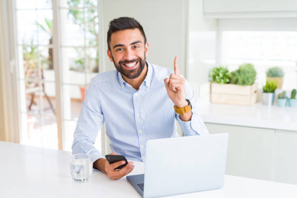 istock Handsome hispanic business man using laptop and smartphone surprised with an idea or question pointing finger with happy face, number one 1167773422