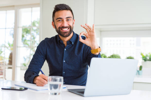 istock Handsome hispanic man working using computer and writing on a paper doing ok sign with fingers, excellent symbol 1167774378