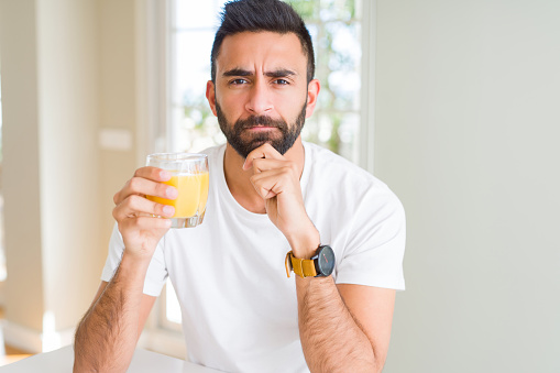 Handsome hispanic man drinking healthy orange juice serious face thinking about question, very confused idea