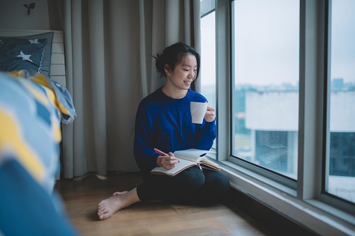 an asian chinese female student sitting beside the bed in bedroom with her coffee cup and writing on the note pad with toothy smile