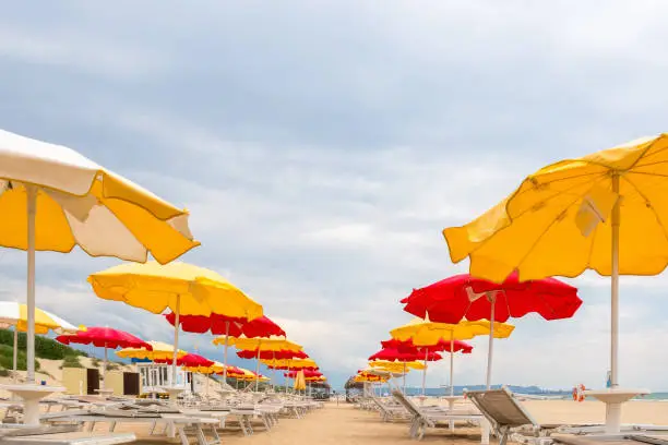 Yellow and red umbrellas and white sunbeds on an empty beach against a cloudy sky