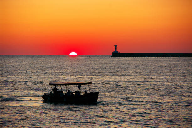 Silhouettes of boat and lighthouse against red sky with partial sun. Sea shore landscape at sunset.