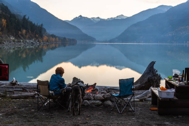 A teen and his dog watch the sun set in front of a campfire on a mountain lake in the fall. On a cool fall evening  teenage boy sits in front of a fire at his camp site enjoying the view with his dog as the sun dips behind the mountains reflecting them in a still Lillooet Lake. pemberton bc stock pictures, royalty-free photos & images