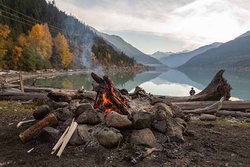 A campfire and kindling sit next to a rock and drift wood covered beach with the mountains and colourful fall leaves reflecting in the calm waters of Lillooet Lake in the background as the sun sets.