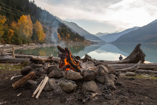 An evening campfire along a mountain lake in the fall. A campfire and kindling sit next to a rock and drift wood covered beach with the mountains and colourful fall leaves reflecting in the calm waters of Lillooet Lake in the background as the sun sets. pemberton bc stock pictures, royalty-free photos & images