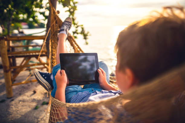 мальчик использует цифровую таблетку, лежащую в гамаке у моря - child beach digital tablet outdoors стоковые фото и изображения