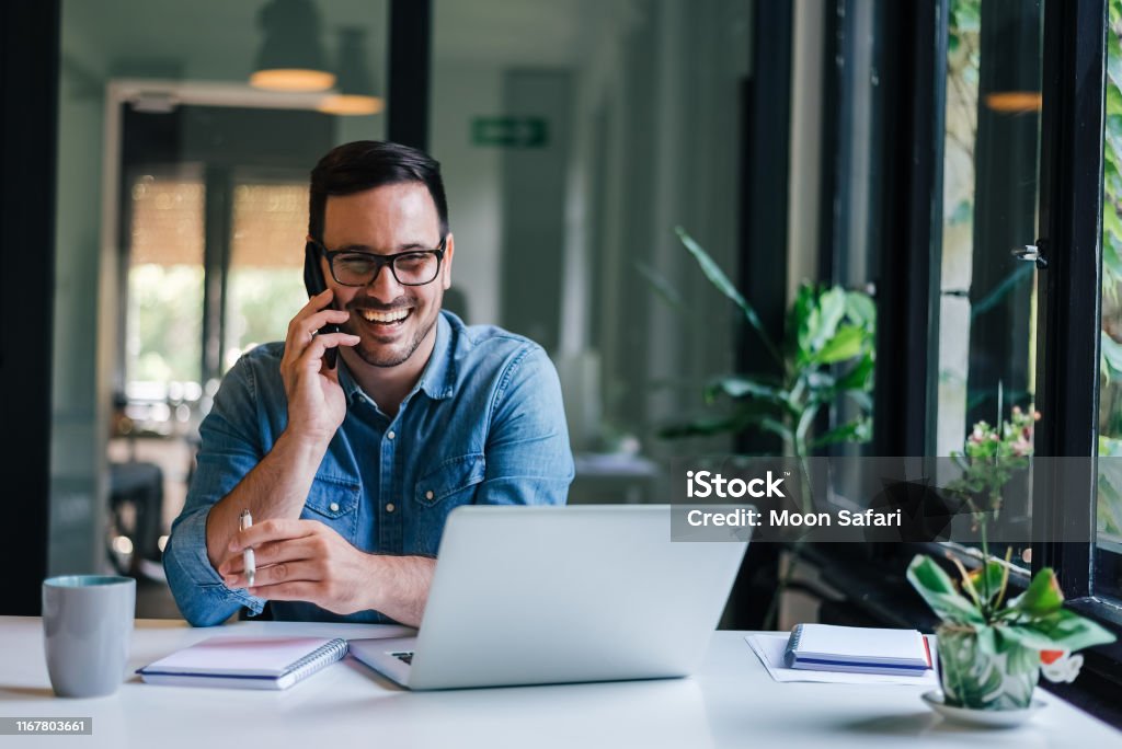 Portrait of young smiling cheerful entrepreneur in casual office making phone call while working with laptop Using Phone Stock Photo
