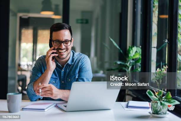 Portrait Of Young Smiling Cheerful Entrepreneur In Casual Office Making Phone Call While Working With Laptop Stock Photo - Download Image Now