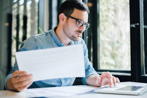 serious pensive thoughtful focused young casual business accountant bookkeeper in office looking at and working with laptop and income tax return papers and documents - computer thinking men people imagens e fotografias de stock