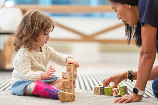 A preschool age girl with a prosthetic leg is at a medical appointment. The child is meeting with her physical therapist. The child is sitting on the floor building with wooden toy blocks. The medical professional is sitting on the floor assisting the girl.