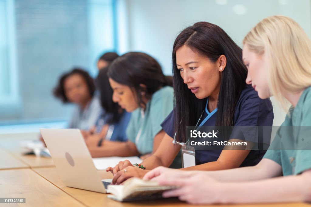 Multi-ethnic group of nursing students in class A multi-ethnic group of female nursing students are attending class together. They are seated at a long table in a classroom. The individuals are working in pairs on a project. The two individuals seated closest to the camera are having a discussion while using a laptop computer. Nurse Stock Photo