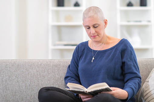 A woman with cancer is undergoing treatment. She has a shaved head because chemotherapy has caused hair loss. The woman is sitting on a couch in her living room at home. She is relaxing while reading a book.