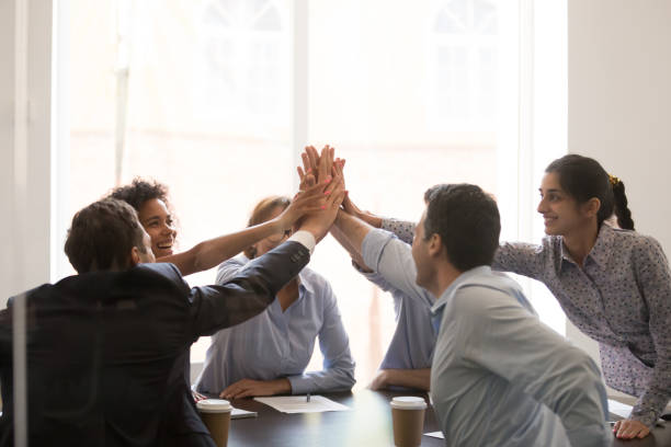 Excited multi racial businesspeople celebrating corporate success giving high five Excited multi racial businesspeople sitting at table conference room achieve corporate success celebrating market leadership giving high five hold hands together feels happy. Team spirit unity concept culture of india stock pictures, royalty-free photos & images