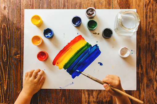 A child holds a brush in his right hand and draws a rainbow on a white sheet of paper.
