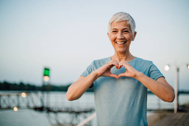 Active senior woman making a heart with her hands Portrait of a happy senior woman making a heart shape with her hands after exercising on the riverbank. body conscious stock pictures, royalty-free photos & images