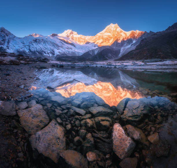istock Snowy mountain with illuminated peaks is reflected in beautiful lake at sunrise. Panoramic landscape with lighted rocks, blue sky, pond, stones in water at dawn. Himalayan mountains in Nepal. Nature 1167856643
