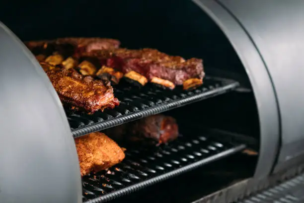 Professional kitchen appliance. Closeup of poultry, beef and pork meat, ribs cooked in BBQ smoker.