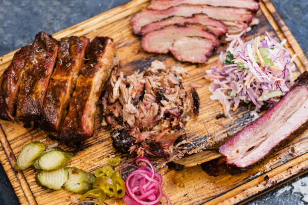 Smoked meat assortment on wooden board. Top view of sliced beef brisket, pulled pork, ribs, coleslaw salad, pickles.