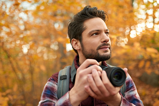Portait of an attractive millennial man walking in autumn nature and doing digital photography with DSLR camera on a path surrounded by orange leaves