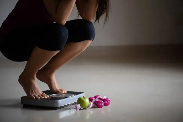 Photo of Close up upset female leg stepping on weigh scales with measuring tape, pink dumbbell and green apple. Healthy lifestyle, food and sport concept.