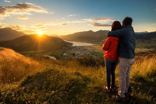 A young couple viewing sunset from the top of a mountain. The beautiful sun produces a warm orange glow to the entire image. Queenstown, New Zealand is a romantic getaway for lovers.