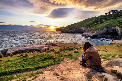 A sad lonely girl sitting on rock watching sunset. She admires ocean view, cliff, rolling hills, sunset, coves, rock formation while reflecting about the meaning of life.