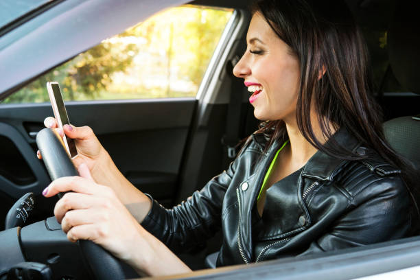 happy smiling woman holding her smart phone sitting inside a car. female driver laughing reading message and chatting in mobile. girl looks at her cellphone screen and smiles while being at wheel. - mirth imagens e fotografias de stock