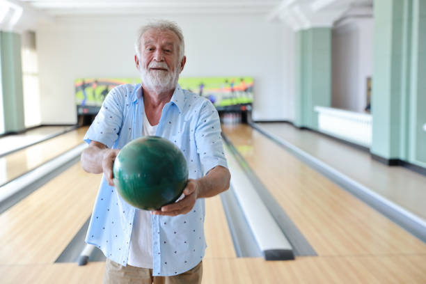 istock Older caucasian men, white beard and white hair in white and blue shirt playing bowling while he holding bowling ball to someone in sport club 1167897930