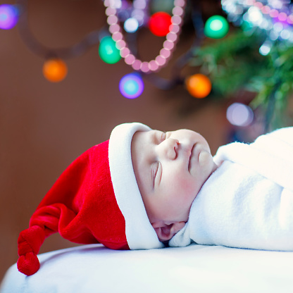 One week old newborn baby in Santa hat wrapped in blanket near Christmas tree with colorful garland lights on background. Closeup of cute child, little baby sleeping. Family, Xmas, birth, new life