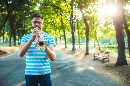Young caucasian man playing trumpet in public park in shade of trees.