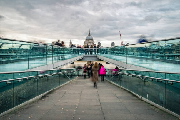 Motion blurred image providing a view of the busy Millennium Bridge with commuters and tourists on Thames Southbank towards the London city skyline with Saint Paul's Cathedral on a cloudy afternoon with moody sky. Shot with Canon EOS full frame system with extra wide lens. The image is ideal for horizontal background with plenty of copy space for your message.