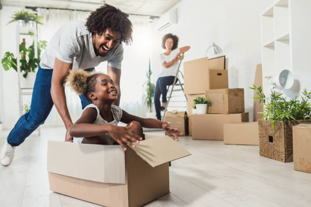 Young Smiling Happy African-american Family Unpacking During Move. New Home. Little preschooler daughter sitting in cardboard box, father rolling her. Smiling mother looking them. unpacking stock pictures, royalty-free photos & images