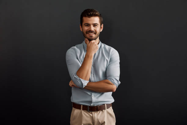 istock Happy to work here. Cheerful young bearded man looking at camera with smile and touching his chin while standing against dark background 1167909256