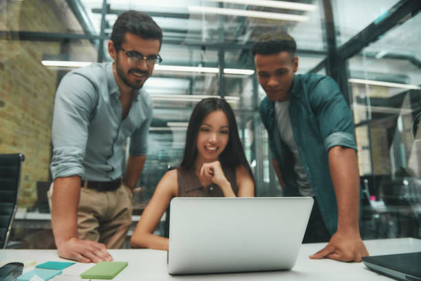 istock Using modern technologies. Group of three young and cheerful employees looking at screen of laptop and smiling while working in modern office 1167910029