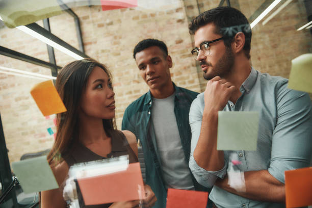 istock IT team. Cheerful male and female colleagues planning working process and using colorful stickers while standing in front of glass wall 1167910062