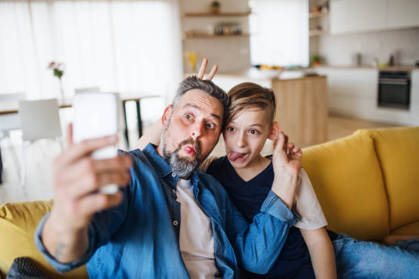 istock Father with small son sitting on sofa indoors, grimacing when taking selfie. 1167921236
