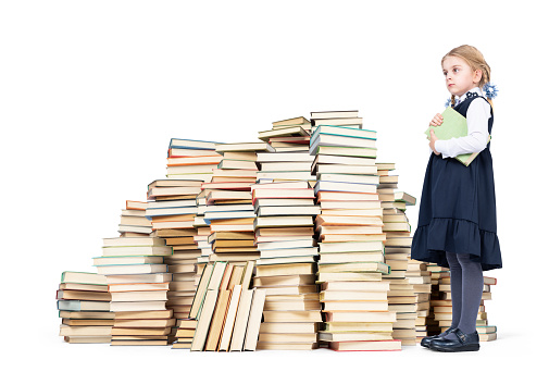 Little confident girl schoolgirl stands next to a huge stack of books and holds a book in her hand. Isolated on white background. Education concept.