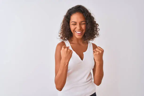 Photo of Young brazilian woman wearing casual t-shirt standing over isolated white background very happy and excited doing winner gesture with arms raised, smiling and screaming for success. Celebration concept.