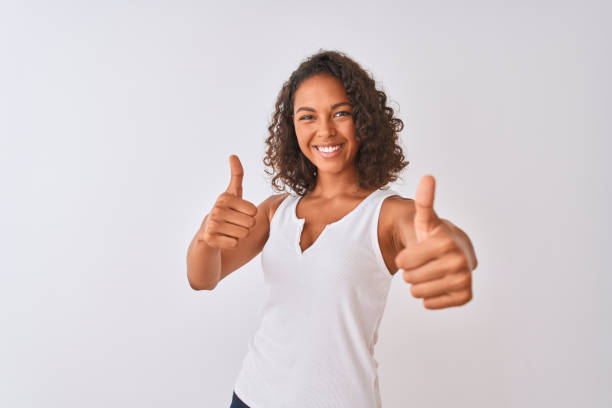 junge brasilianerin trägt lässiges t-shirt, das über einem isolierten weißen hintergrund steht und eine positive geste mit der hand, die daumen hoch lächelnd und glücklich über den erfolg genehmigt. gewinner geste. - thumbs up human thumb human finger brown hair stock-fotos und bilder