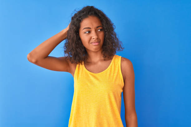 mulher brasileira nova que desgasta o t-shirt amarelo que está sobre o fundo azul isolado confunda e maravilha sobre a pergunta. incerto com dúvida, pensando com a mão na cabeça. conceito pensativo. - teenage girls ideas blue yellow - fotografias e filmes do acervo