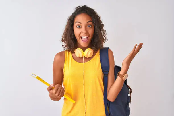 Photo of Brazilian student woman wearing backpack holding notebook over isolated white background very happy and excited, winner expression celebrating victory screaming with big smile and raised hands