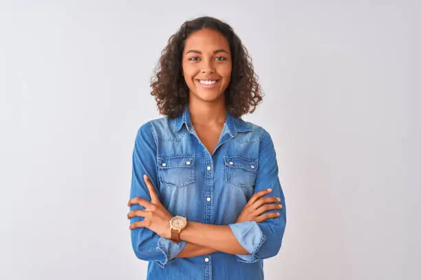 Photo of Young brazilian woman wearing denim shirt standing over isolated white background happy face smiling with crossed arms looking at the camera. Positive person.