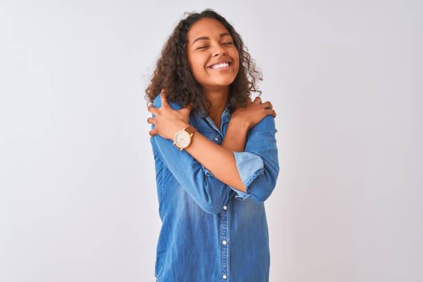 joven mujer brasileña con camisa de mezclilla de pie sobre fondo blanco aislado abrazándose feliz y positivo, sonriendo confiado. amor propio y autocuidado - photography teenagers only vertical teenager fotografías e imágenes de stock