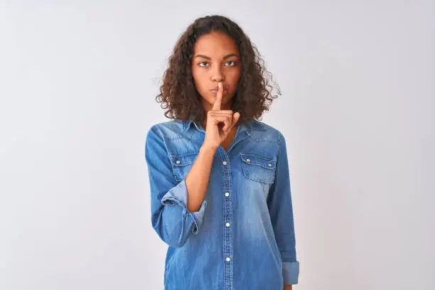 Photo of Young brazilian woman wearing denim shirt standing over isolated white background asking to be quiet with finger on lips. Silence and secret concept.