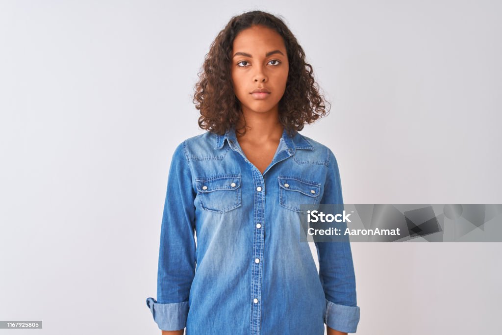 Young brazilian woman wearing denim shirt standing over isolated white background Relaxed with serious expression on face. Simple and natural looking at the camera. Serious Stock Photo